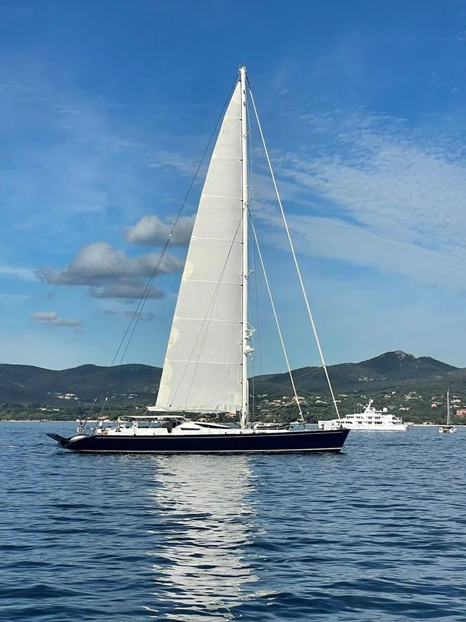 A sleek sailing yacht glides gracefully on calm waters against a backdrop of rolling hills and a clear blue sky. aboard MUMU Yacht for Sale