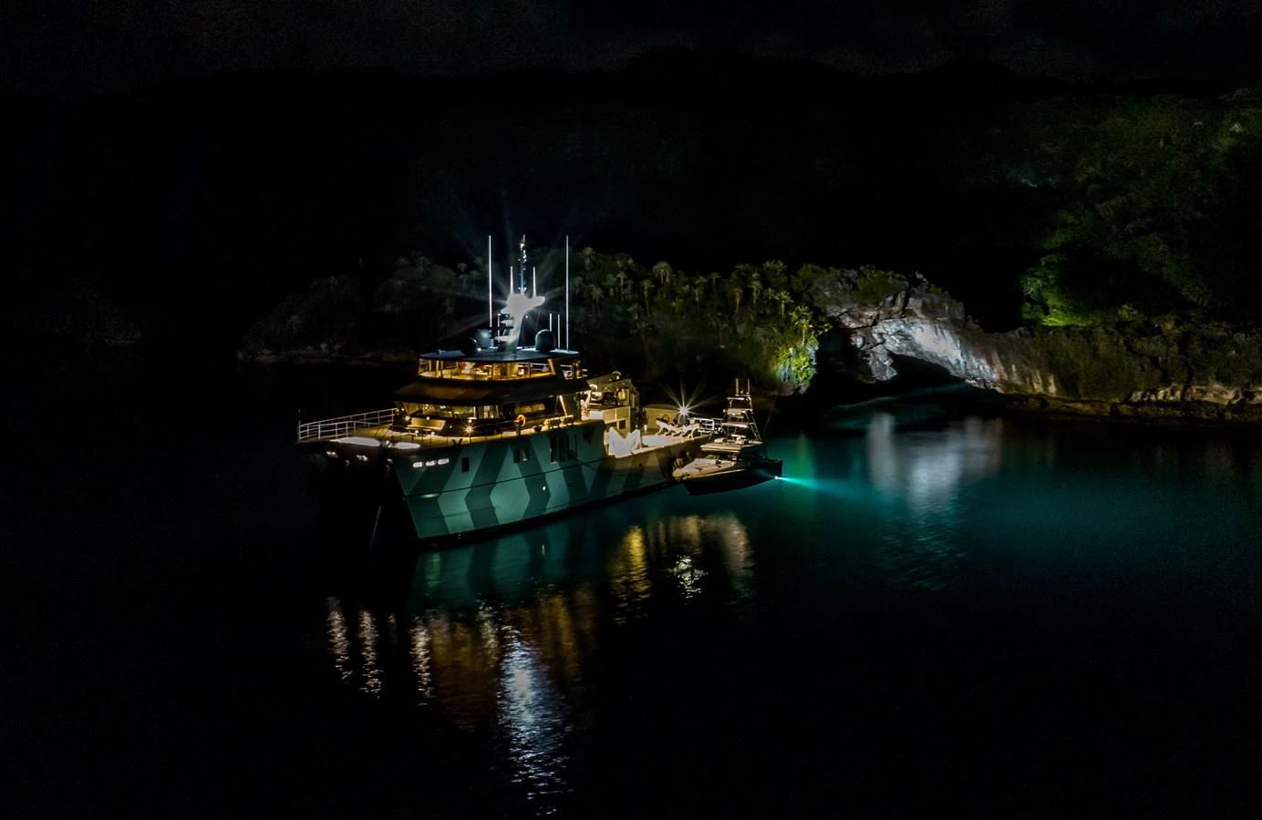 The yacht is beautifully illuminated at night, anchored in a serene cove with a cave in the background. aboard THE BEAST Yacht for Sale