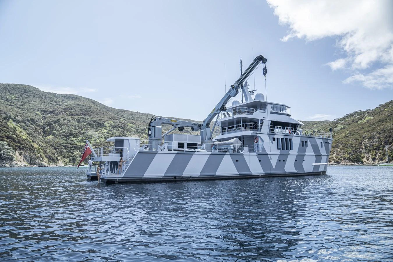 A robust yacht with a distinctive camouflage pattern is anchored in a serene bay surrounded by lush hills. aboard THE BEAST Yacht for Sale