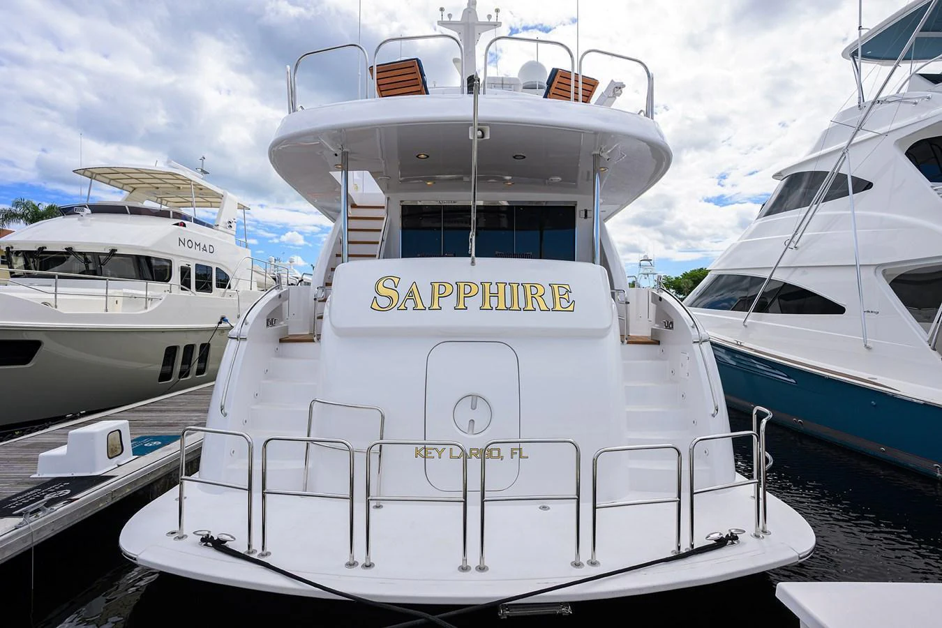 The yacht is docked at a marina, showcasing its elegant stern with a clean, white finish and gold lettering. aboard SAPPHIRE Yacht for Sale