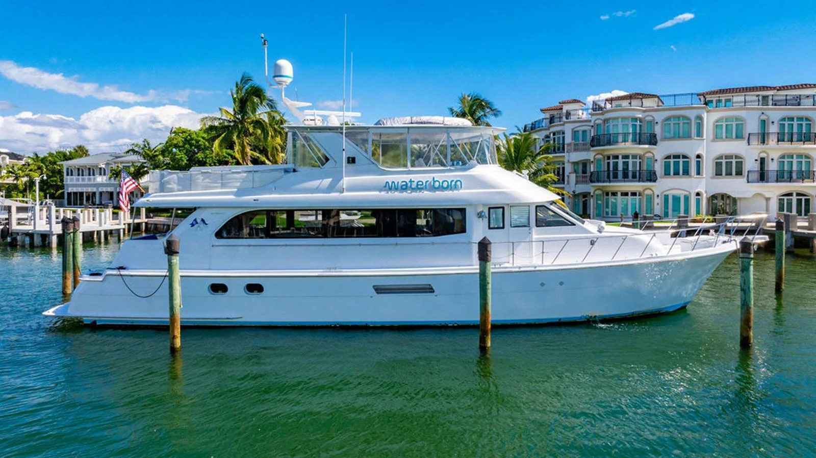A sleek white yacht is docked in a picturesque marina with luxurious waterfront homes in the background. aboard WATER BORN Yacht for Sale