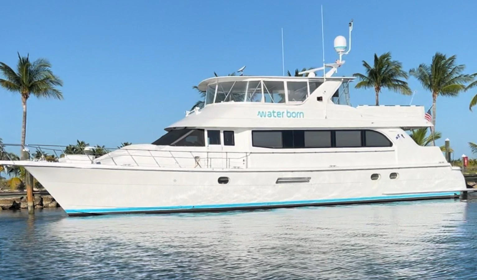 A sleek white yacht is docked in a tropical marina, showcasing its elegant profile against a backdrop of palm trees and clear blue skies. aboard WATER BORN Yacht for Sale
