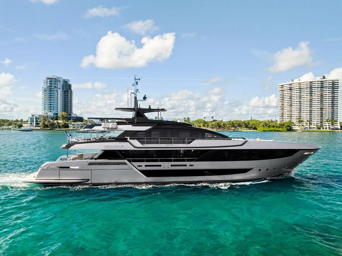 A sleek and modern yacht cruises through clear blue waters with a cityscape in the background. aboard TENACIOUS Yacht for Sale