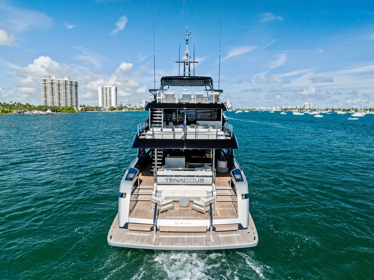 The yacht is seen from the rear, showcasing its spacious aft deck and sleek design as it cruises on the water. aboard TENACIOUS Yacht for Sale