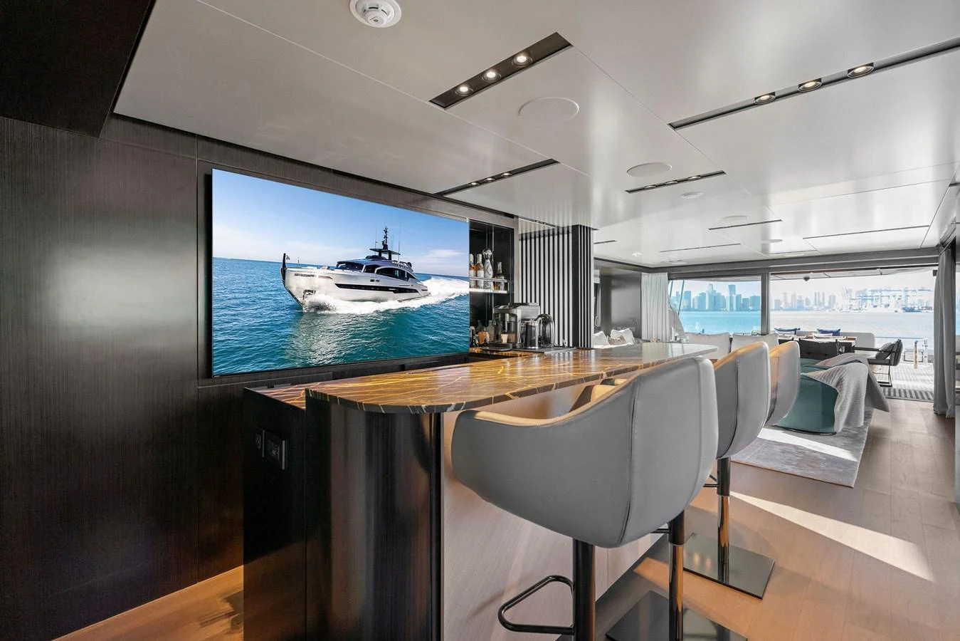 The luxurious salon features a sleek bar area with modern seating and a large TV displaying a yacht on the water. aboard CAFFEINE Yacht for Sale