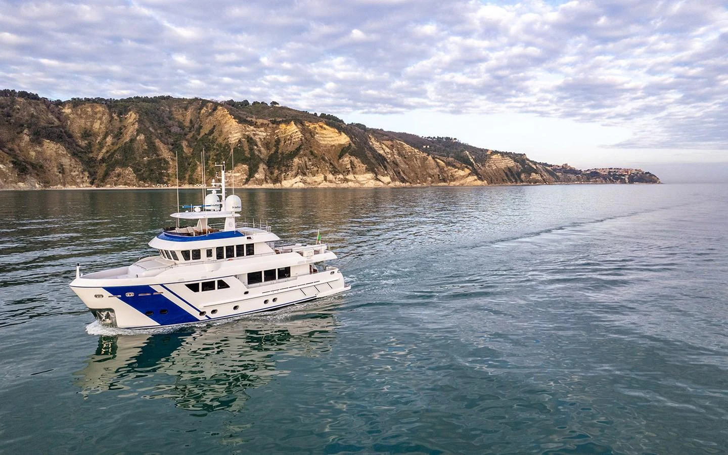 A sleek yacht cruises along a serene coastline under a partly cloudy sky. aboard EMPIRE Yacht for Sale