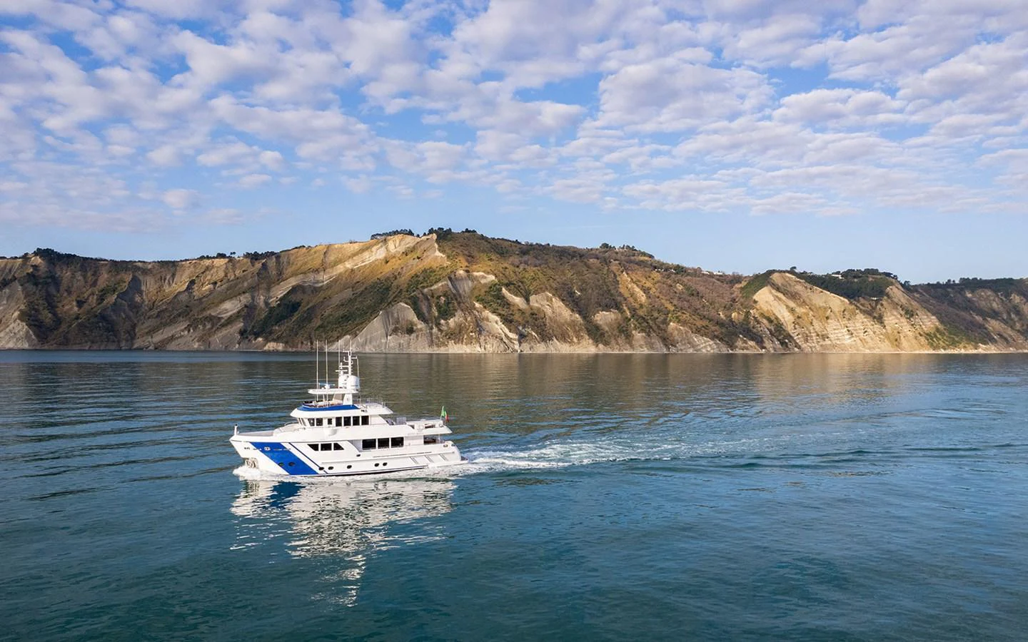A sleek yacht cruises along a serene coastline under a partly cloudy sky. aboard EMPIRE Yacht for Sale