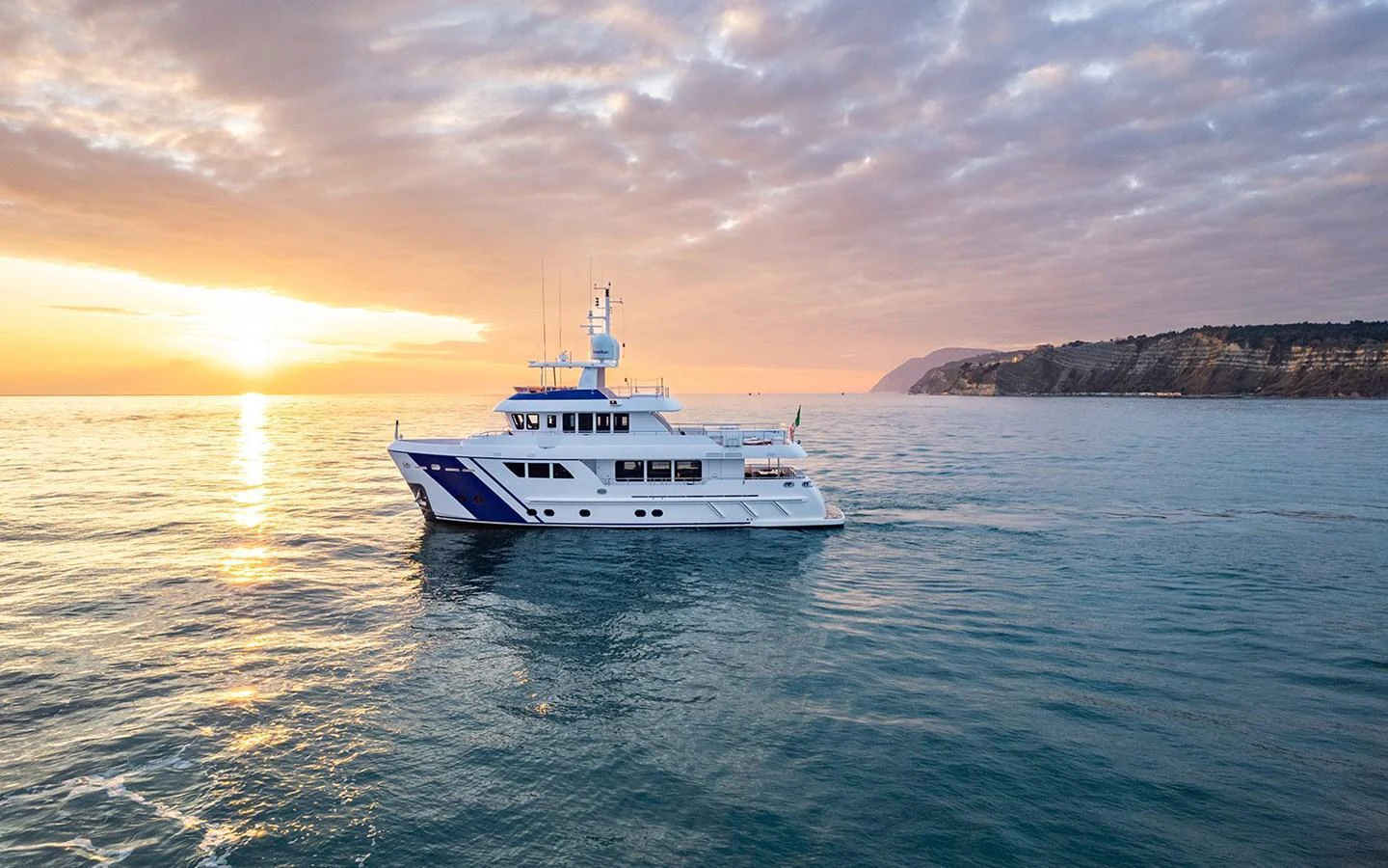 A luxurious yacht cruises serenely on calm waters at sunset, with a picturesque coastline in the background. aboard EMPIRE Yacht for Sale