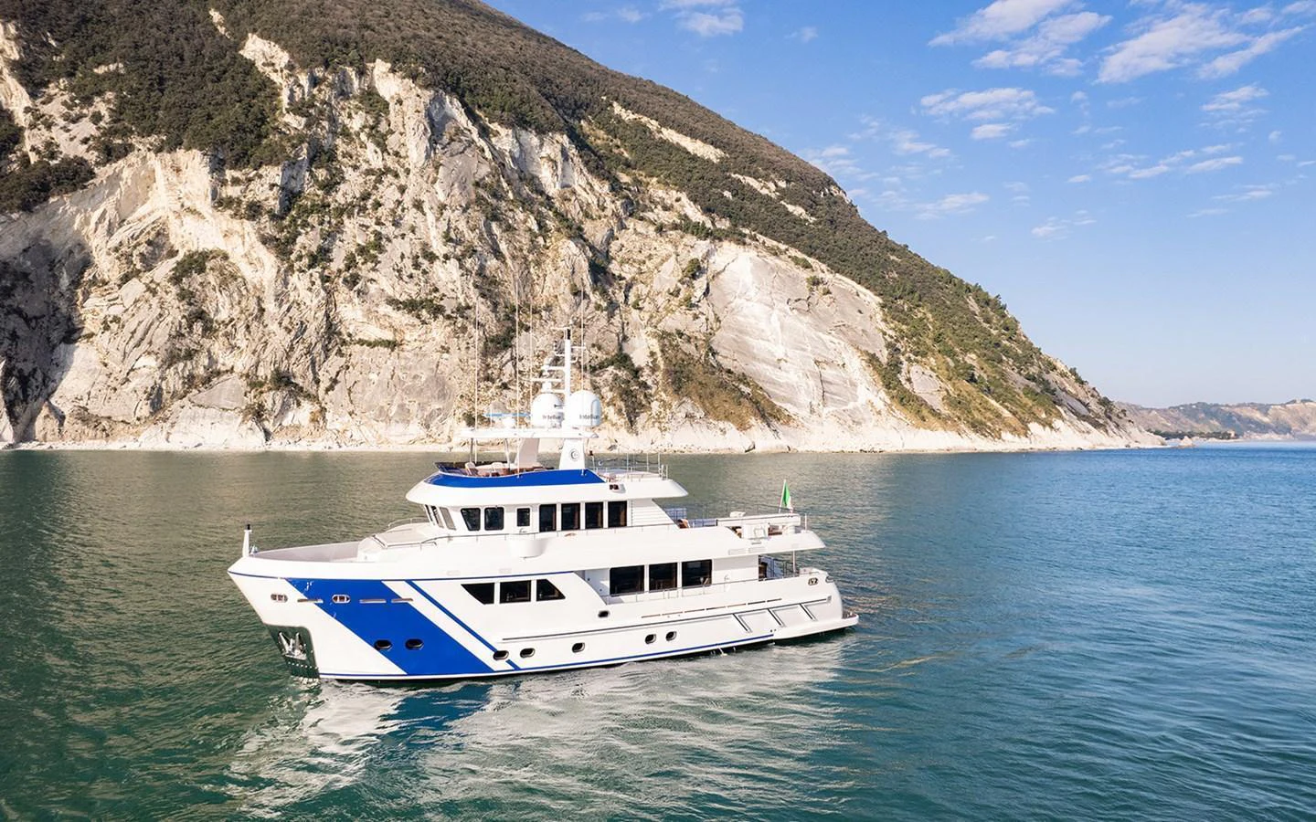 A sleek yacht cruises along a scenic coastline with clear blue waters and rugged cliffs in the background. aboard EMPIRE Yacht for Sale