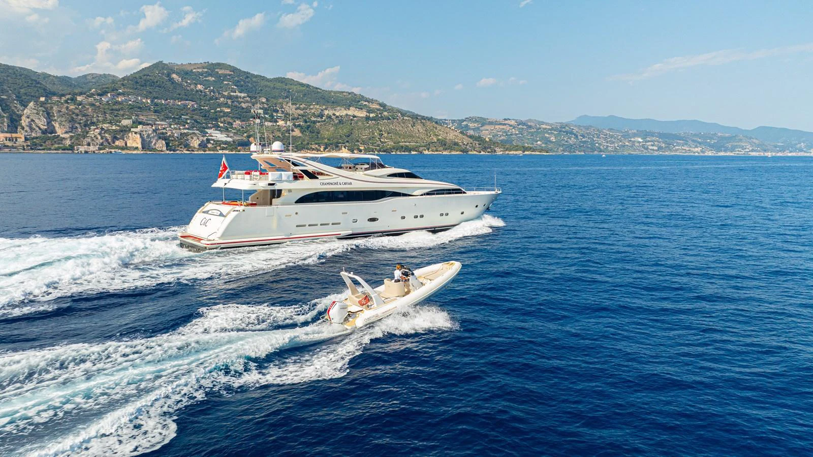 A sleek yacht cruises along the coastline with a tender boat following closely behind. aboard CHAMPAGNE AND CAVIAR Yacht for Sale