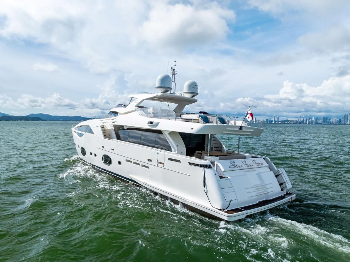 A sleek and modern yacht cruises through calm waters with a cityscape in the background. aboard SHA SHA SHA IV Yacht for Sale