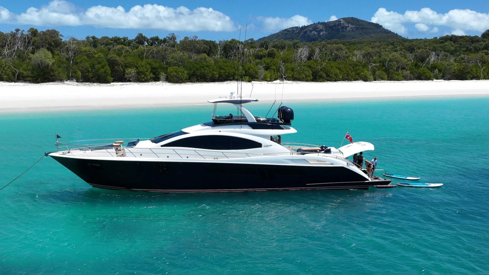 A sleek yacht is anchored in crystal-clear turquoise waters near a pristine beach with lush greenery in the background. aboard LA MAR Yacht for Sale