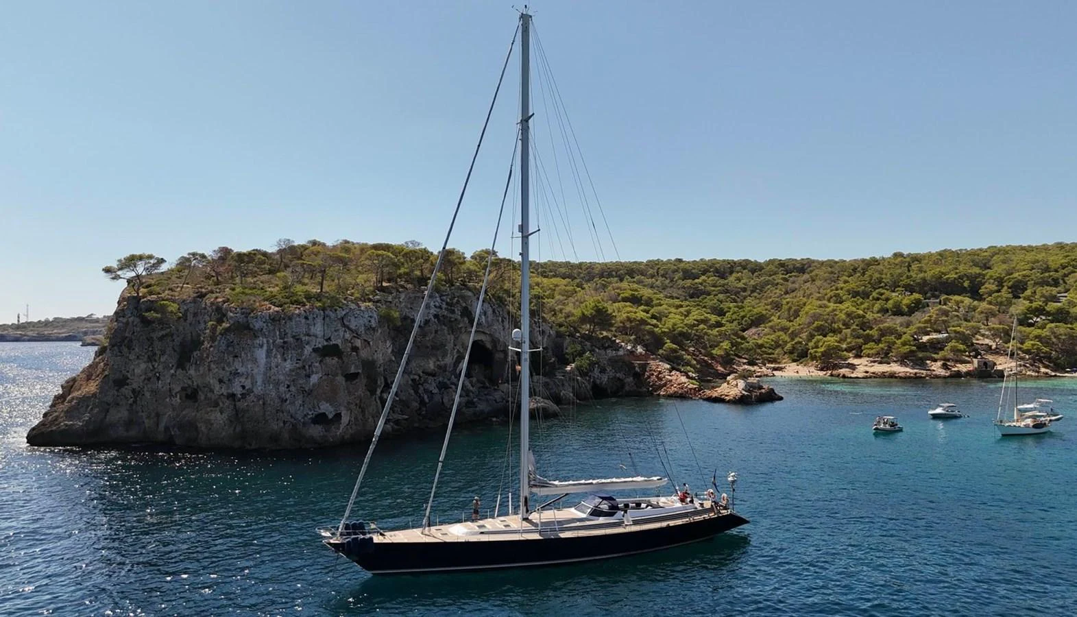 A sleek sailing yacht glides gracefully on serene blue waters near a rocky, tree-covered coastline. aboard PERSISTENCE Yacht for Sale