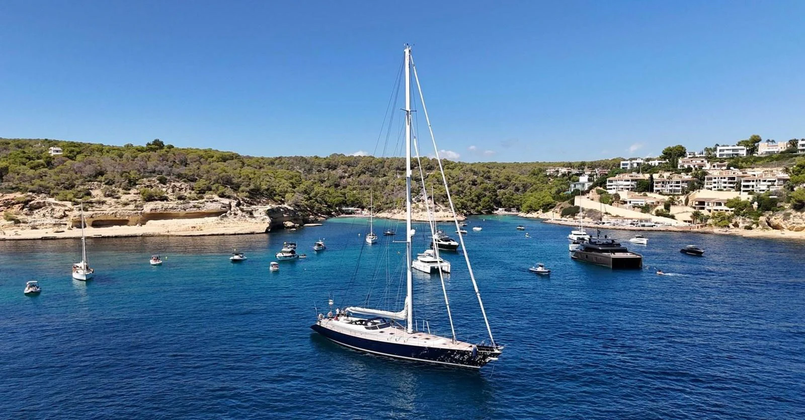 A sleek sailing yacht is anchored in a serene bay surrounded by other boats and a picturesque coastline. aboard PERSISTENCE Yacht for Sale
