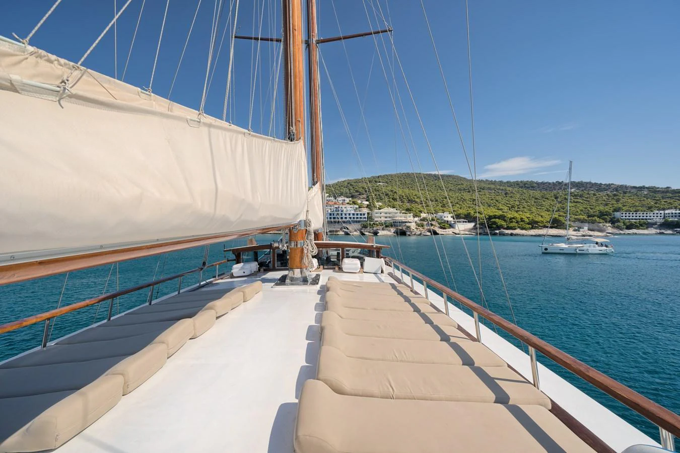 The foredeck of this yacht features ample sunbathing space with comfortable cushions, set against a stunning coastal backdrop. aboard THALASSA Yacht for Sale