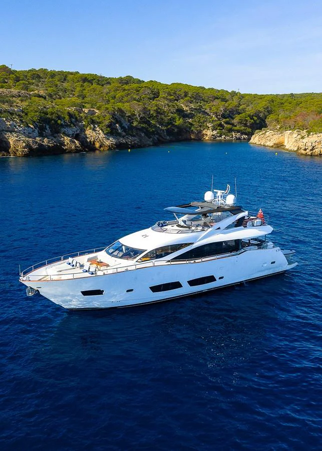A sleek white yacht cruises through tranquil blue waters near a rocky coastline with lush greenery. aboard THIS TIME NEXT YEAR Yacht for Sale