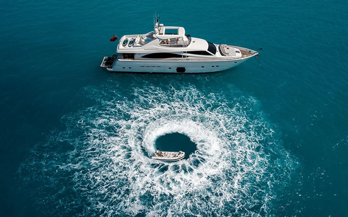 A luxurious yacht is anchored in clear blue waters with a tender creating a circular wake nearby. aboard ONYX Yacht for Sale