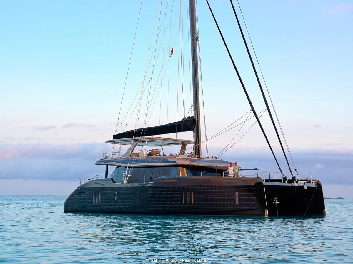 A sleek, modern catamaran yacht glides gracefully on calm waters under a clear sky. aboard THE IDLER Yacht for Sale