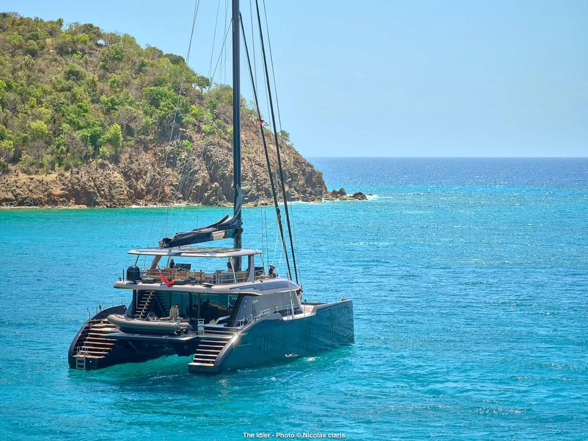 A sleek catamaran yacht is anchored in a serene turquoise bay with lush green hills in the background. aboard THE IDLER Yacht for Sale
