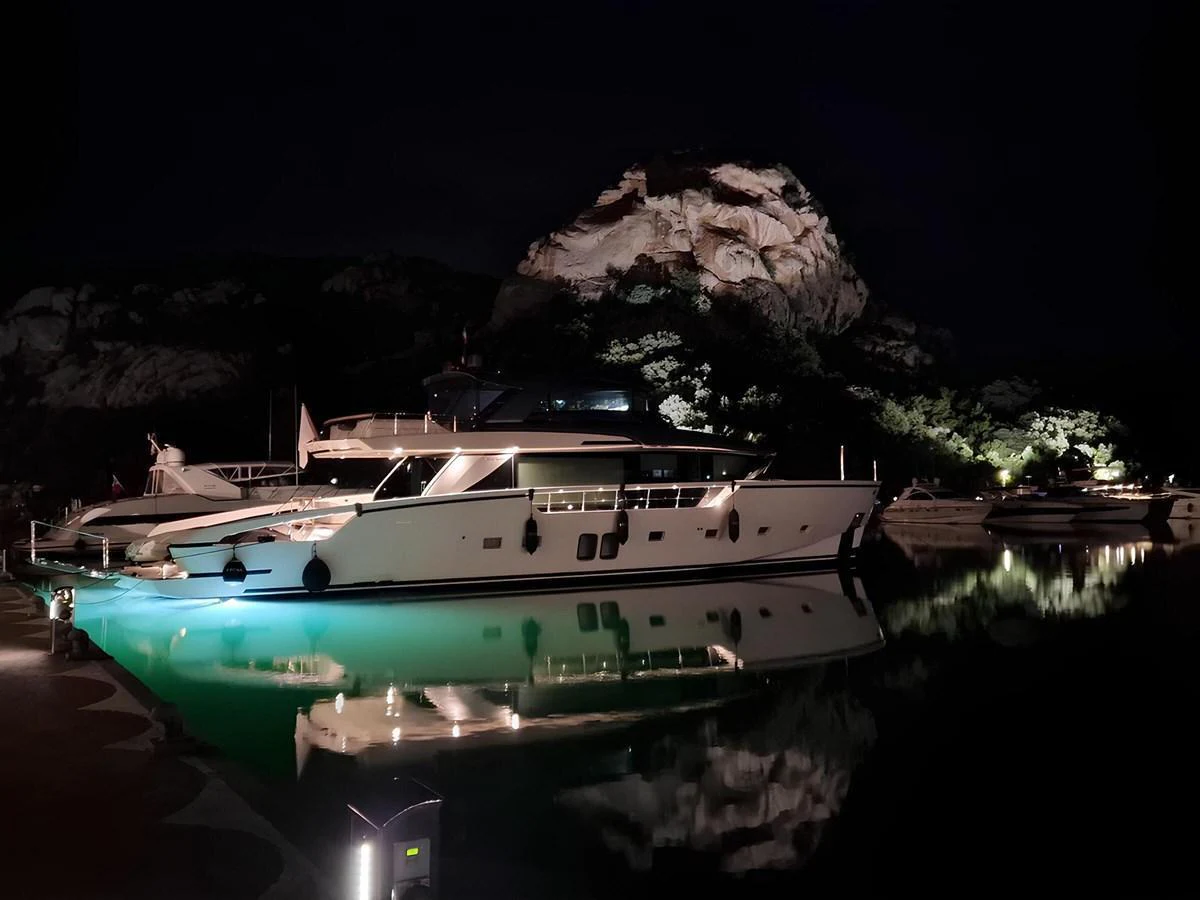 A sleek yacht is beautifully illuminated at night, docked in a serene marina with a rocky backdrop. aboard LYDIA Yacht for Sale
