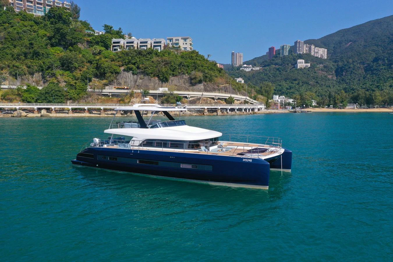 A sleek and modern yacht is anchored in a serene coastal area with lush greenery and buildings in the background. aboard LAGOON SIXTY 7 Yacht for Sale