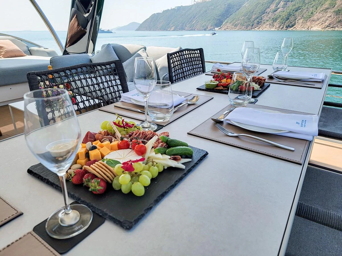 The yacht's dining area is elegantly set with a variety of fresh fruits, cheeses, and gourmet appetizers, offering a luxurious alfresco dining experience with a scenic view. aboard LAGOON SIXTY 7 Yacht for Sale