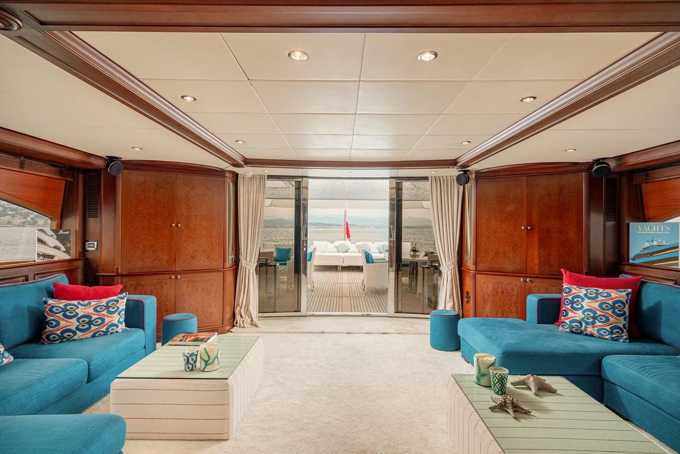The salon features plush blue seating and wooden accents, opening up to a view of the deck and sea. aboard BANG! Yacht for Sale