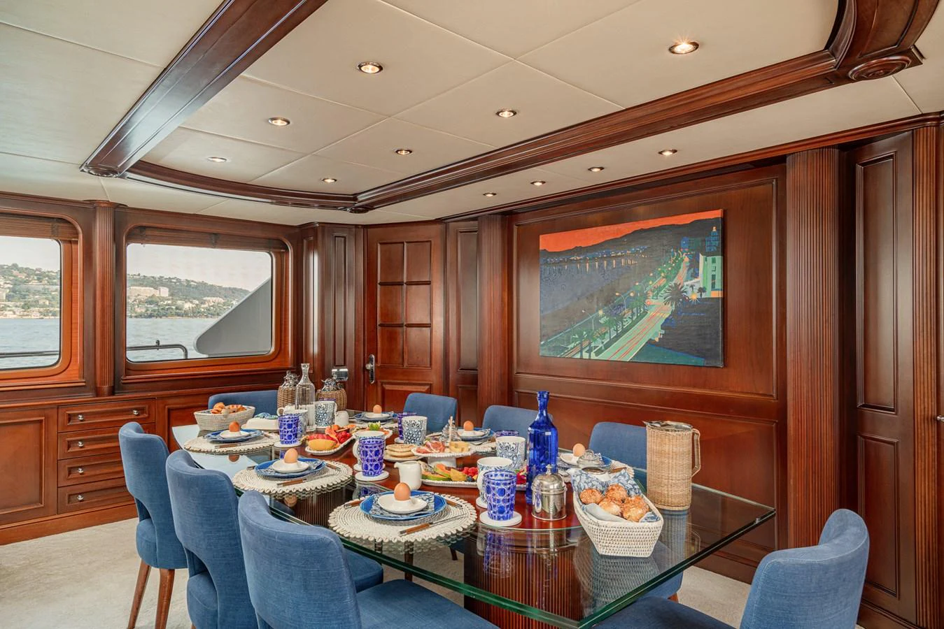 The yacht's dining area features a glass table set for a meal, surrounded by blue upholstered chairs and rich wooden paneling. aboard BANG! Yacht for Sale