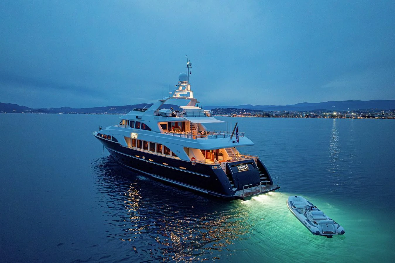 A luxurious yacht is anchored in calm waters during twilight, with its lights illuminating the surroundings. aboard BANG! Yacht for Sale