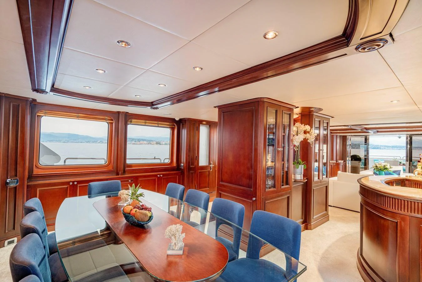 The yacht's dining area features elegant wood paneling, a glass-top table, and plush blue chairs, offering a luxurious setting with scenic views. aboard BANG! Yacht for Sale