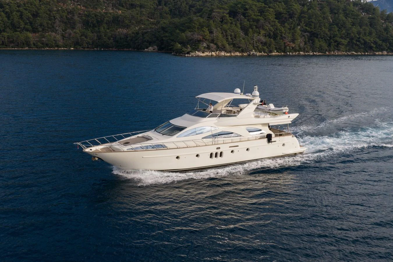 A sleek, luxurious yacht cruises through calm waters with a scenic forested coastline in the background. aboard SENIOR S Yacht for Sale