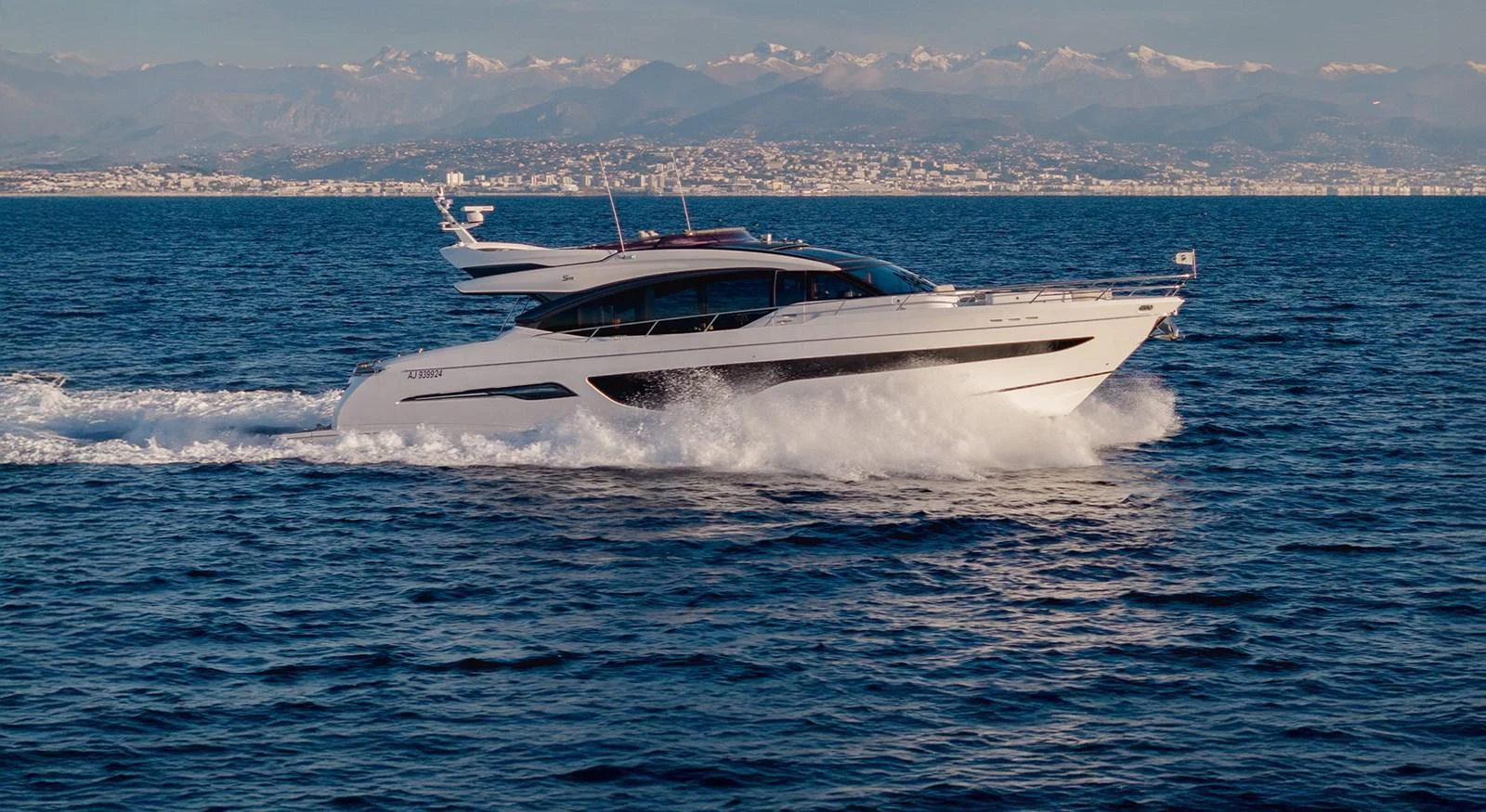 A sleek and modern yacht cruises swiftly across the open sea with a cityscape and mountains in the background. aboard CAMILLE Yacht for Sale