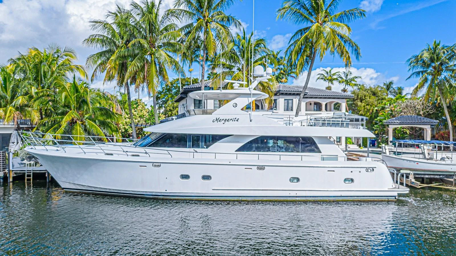 A sleek white yacht is docked in a tropical marina, surrounded by palm trees and waterfront homes. aboard MARGARITA Yacht for Sale