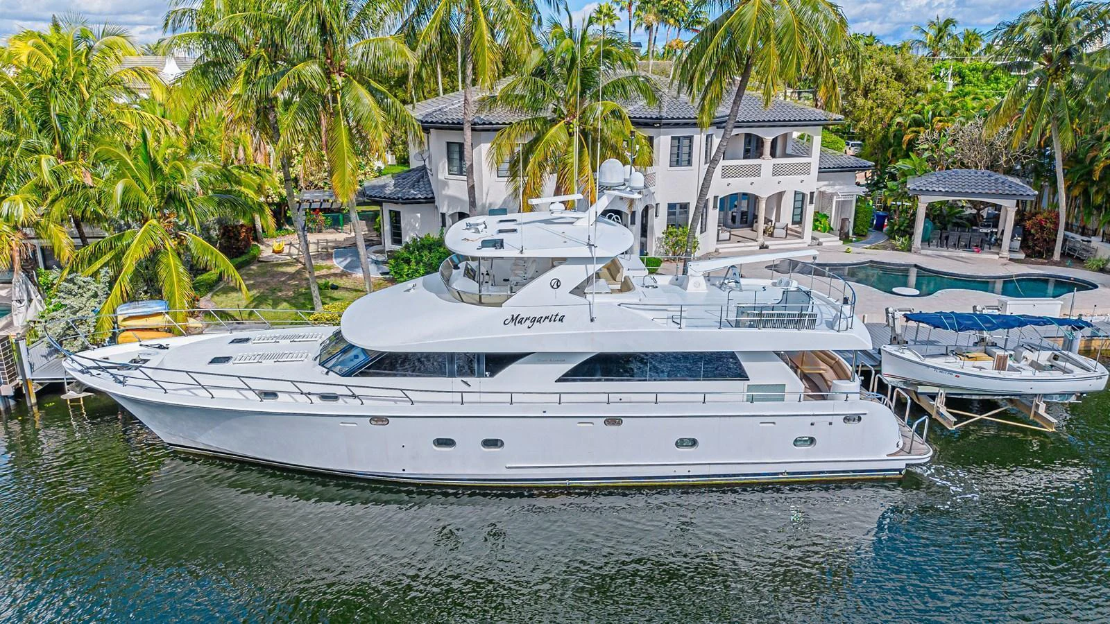 A sleek white yacht is docked beside a luxurious waterfront property, surrounded by lush palm trees. aboard MARGARITA Yacht for Sale