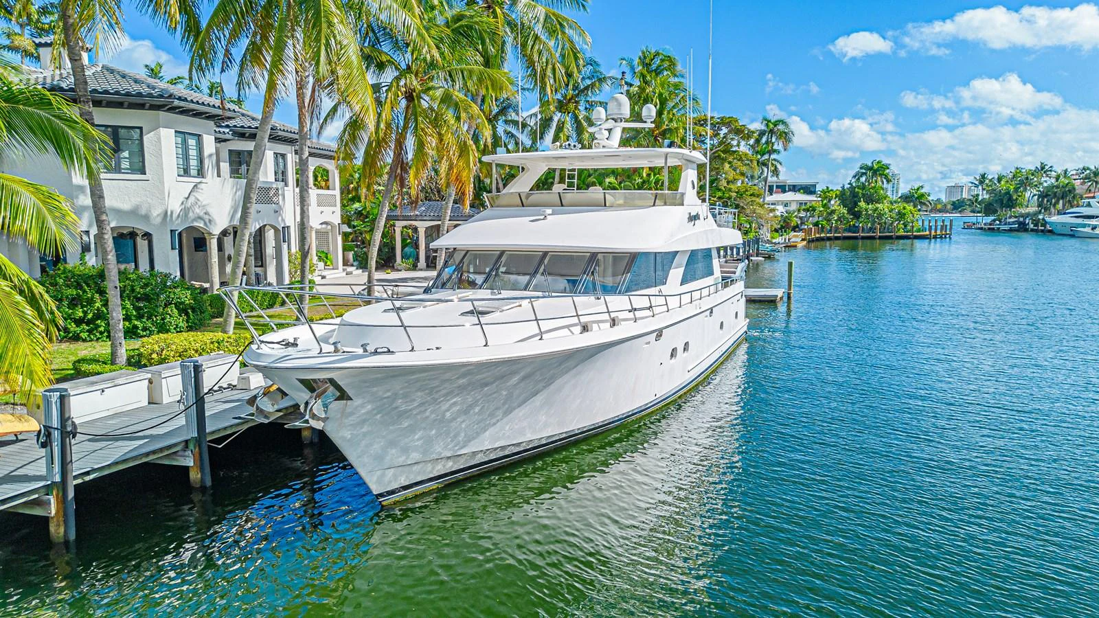 A sleek white yacht is docked in a serene waterfront setting with palm trees and luxurious homes in the background. aboard MARGARITA Yacht for Sale