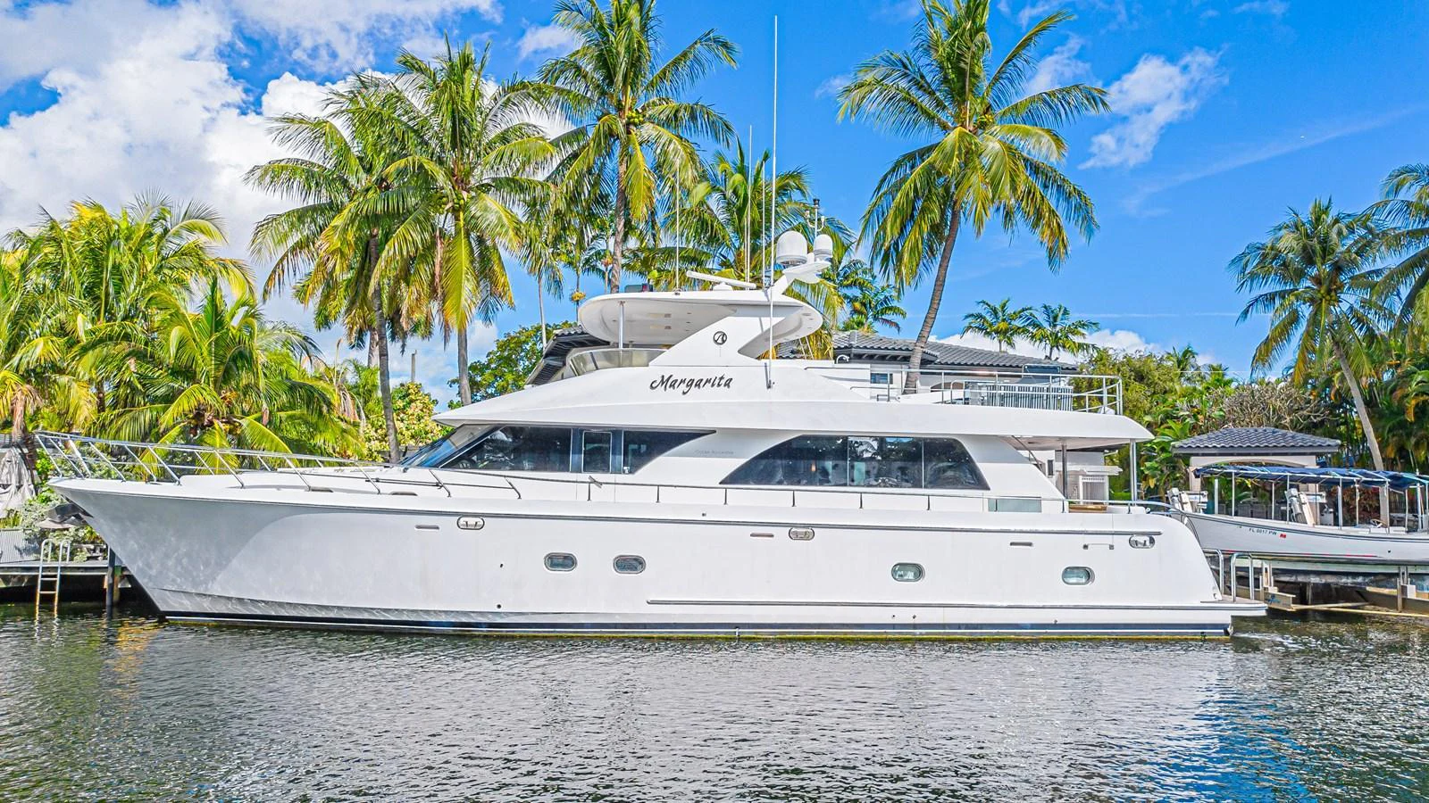 A sleek white yacht is docked beside palm trees under a bright blue sky. aboard MARGARITA Yacht for Sale
