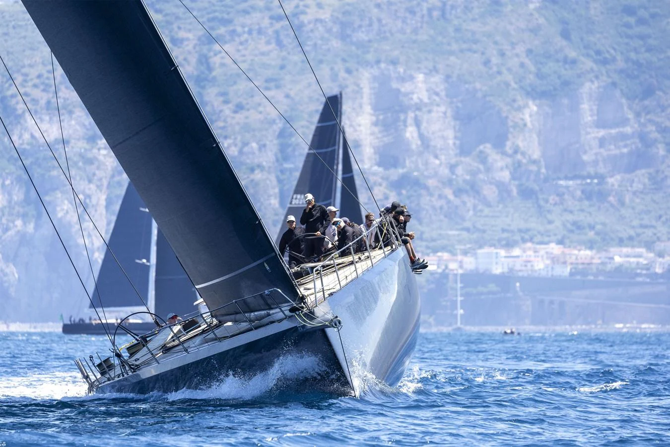 A sleek sailing yacht cuts through the water with its crew on deck, set against a scenic coastal backdrop. aboard BULLITT Yacht for Sale