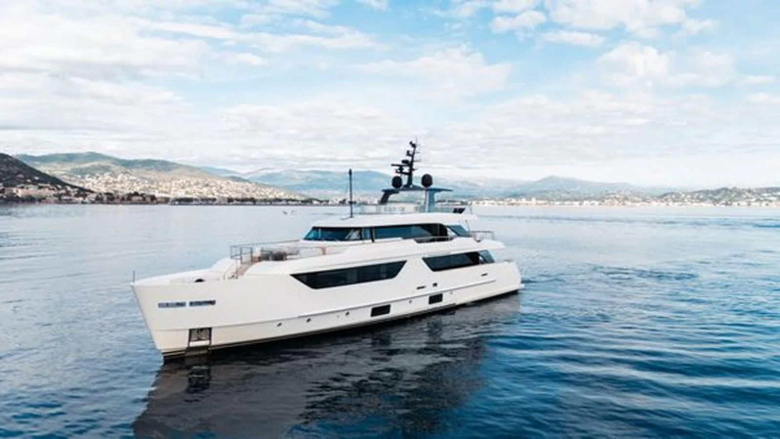 A sleek yacht glides through calm waters with a scenic backdrop of mountains and cityscape. aboard 2022 SANLORENZO SD118 Yacht for Sale
