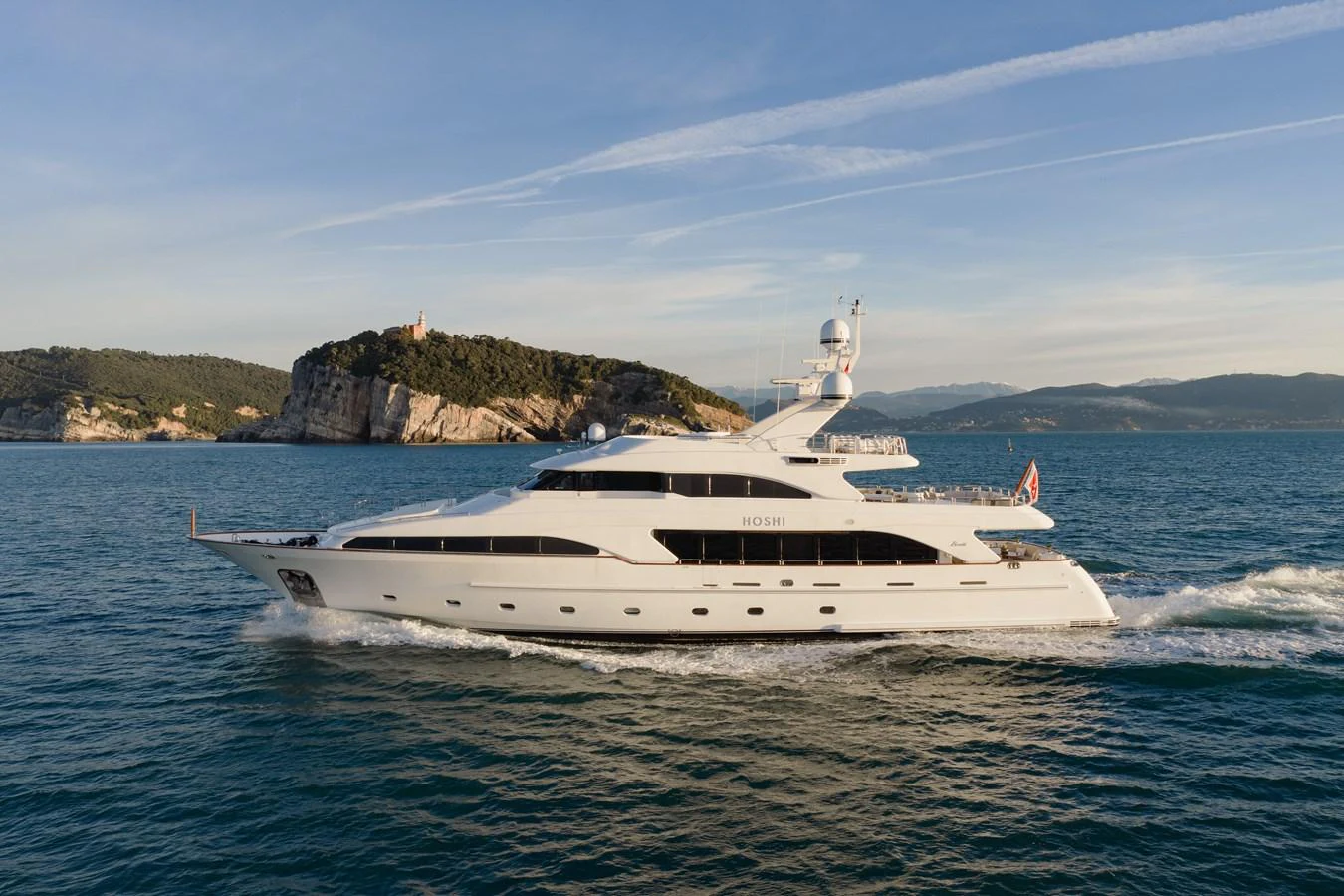 A sleek, white yacht cruises through calm waters with a scenic coastal backdrop. aboard HOSHI Yacht for Sale