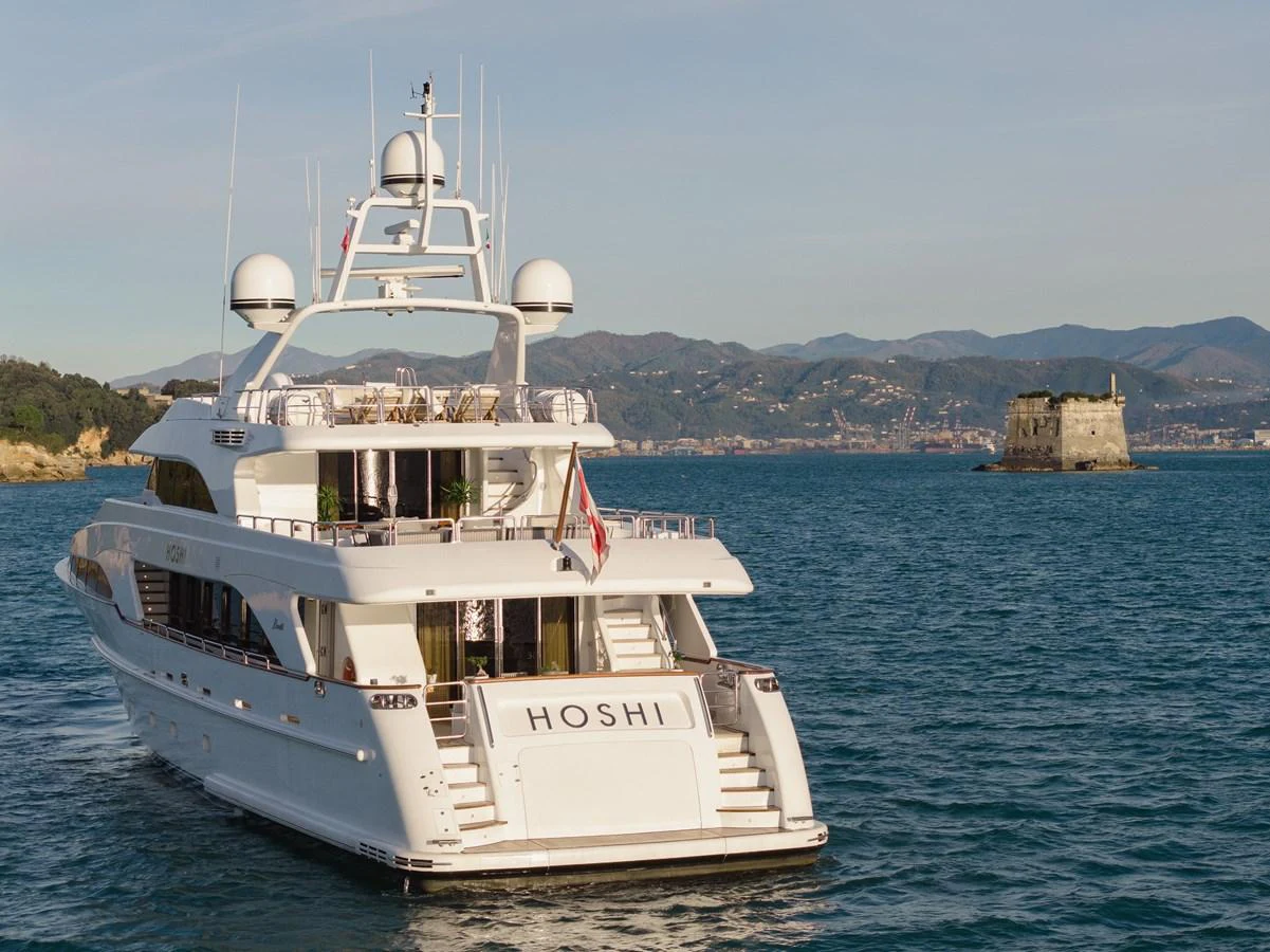 A luxurious yacht is anchored in a serene coastal area with scenic views of the mountains and a historic fort in the background. aboard HOSHI Yacht for Sale