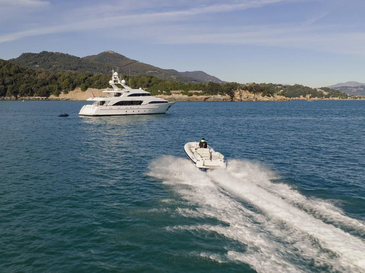 A sleek yacht is anchored in a serene bay with a tender speeding away. aboard HOSHI Yacht for Sale