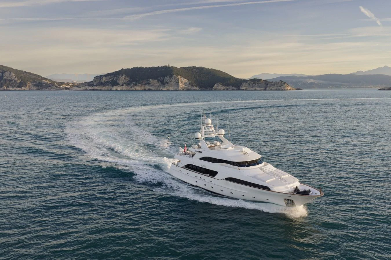 A luxurious yacht cruises through serene waters with picturesque hills in the background. aboard HOSHI Yacht for Sale