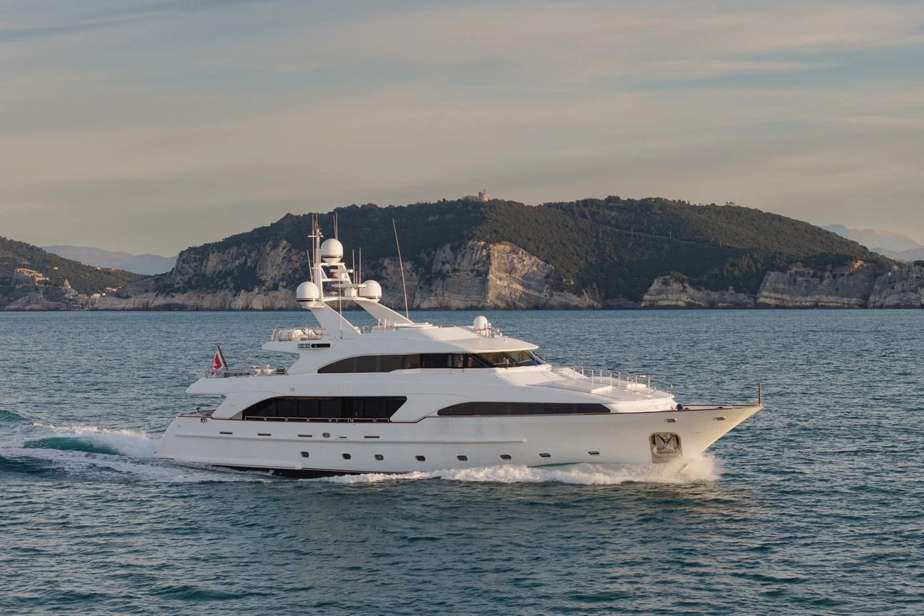 A sleek white yacht cruises through calm waters against a backdrop of scenic hills. aboard HOSHI Yacht for Sale