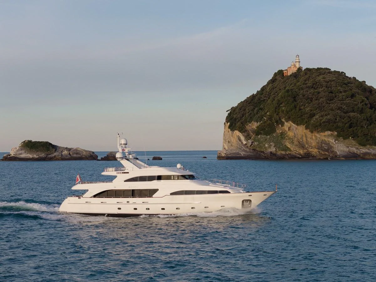 A sleek white yacht cruises through calm blue waters with a scenic island in the background. aboard HOSHI Yacht for Sale