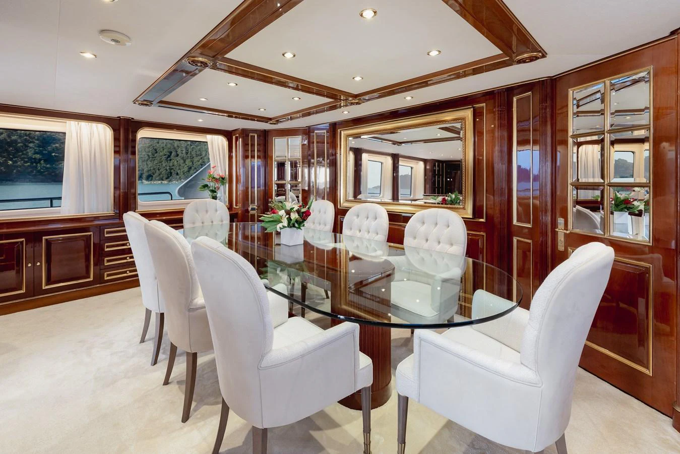 The yacht's dining area features an elegant glass table surrounded by plush white chairs, set against rich wooden paneling and large windows with scenic views. aboard HOSHI Yacht for Sale