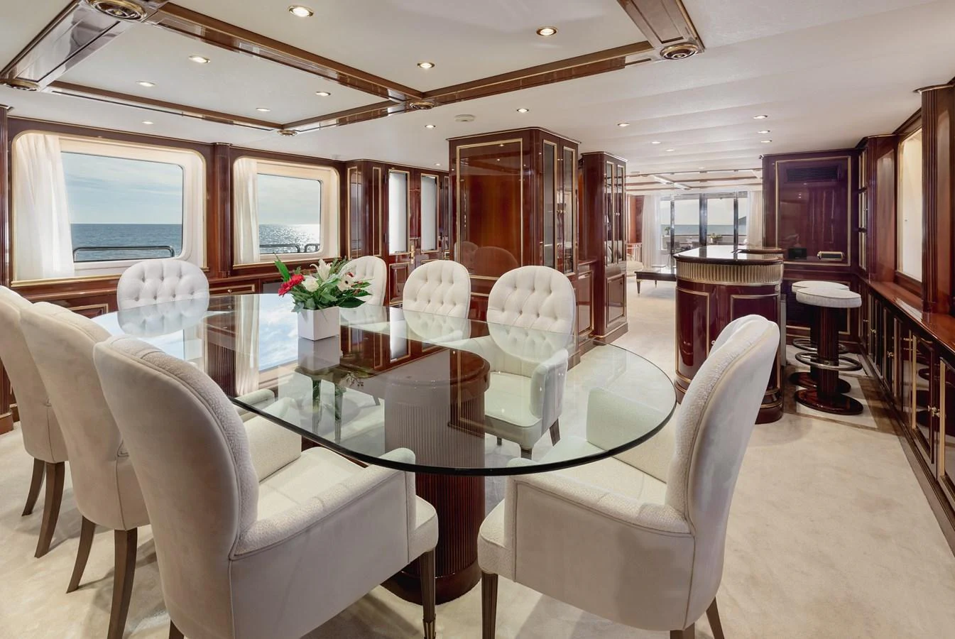 The yacht's dining area features a glass table surrounded by plush white chairs, set against a backdrop of rich wood paneling and large windows offering ocean views. aboard HOSHI Yacht for Sale