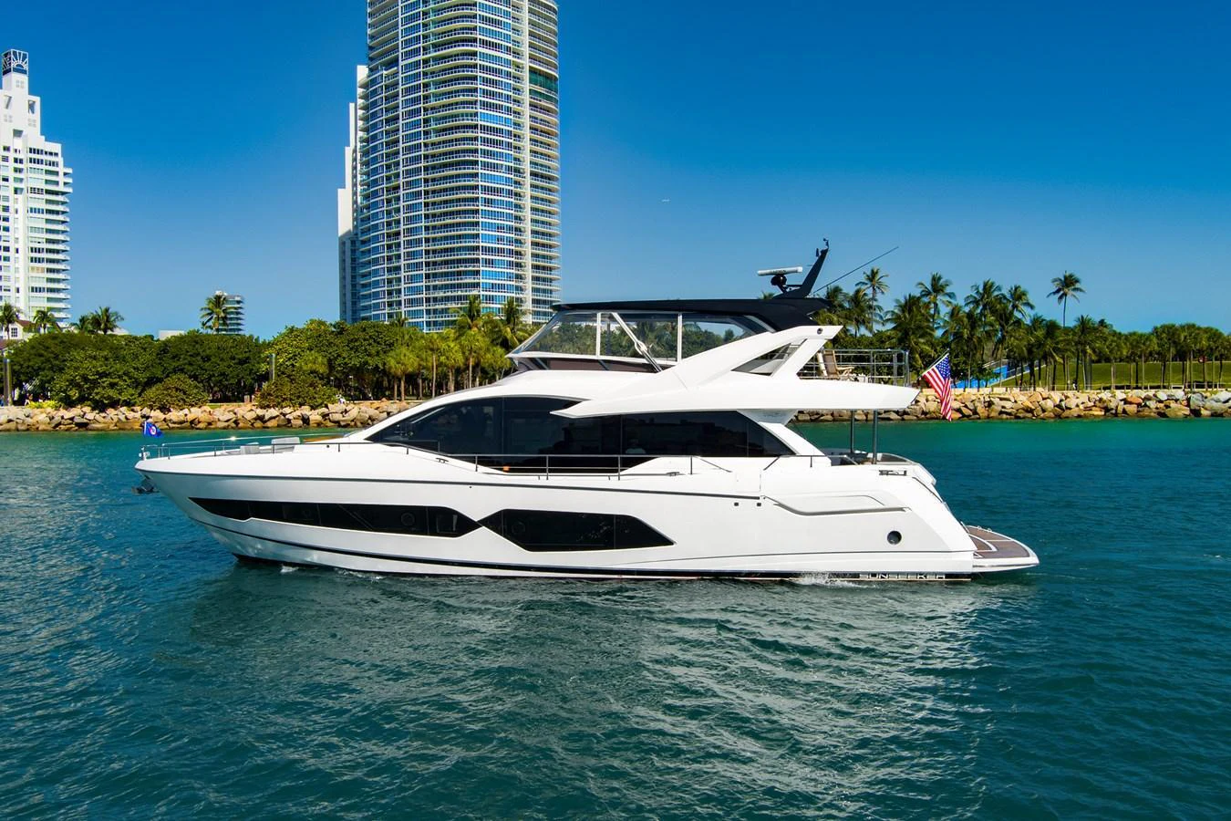 A sleek white yacht cruises along the coastline with modern buildings and palm trees in the background. aboard MISTY K Yacht for Sale