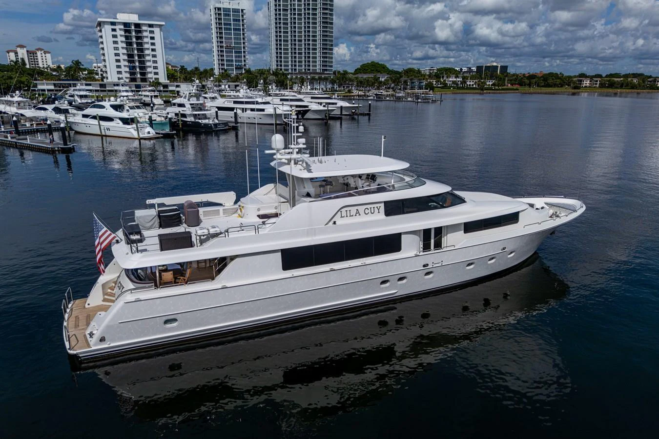 A sleek white yacht is docked in a marina with a cityscape in the background. aboard LILA CUY Yacht for Sale