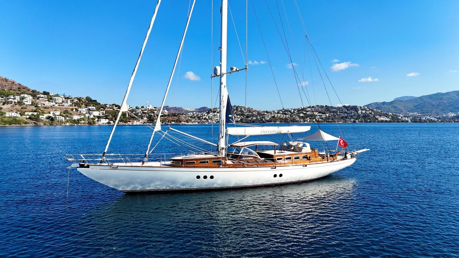 A sleek sailing yacht glides gracefully on calm blue waters with a scenic coastal backdrop. aboard PORTOBELLO Yacht for Sale