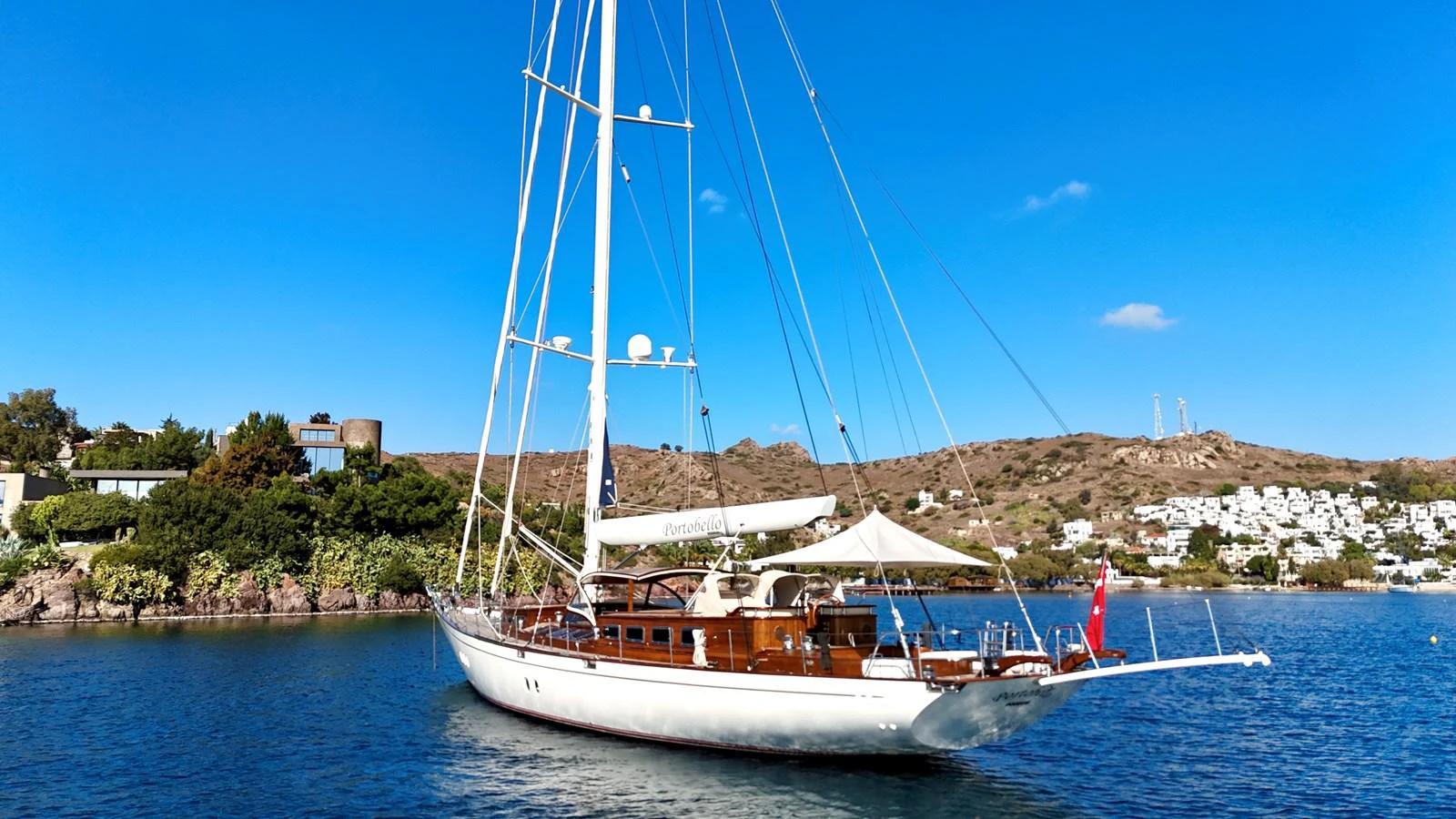 A classic sailing yacht is anchored in a serene bay with a backdrop of hills and clear blue skies. aboard PORTOBELLO Yacht for Sale